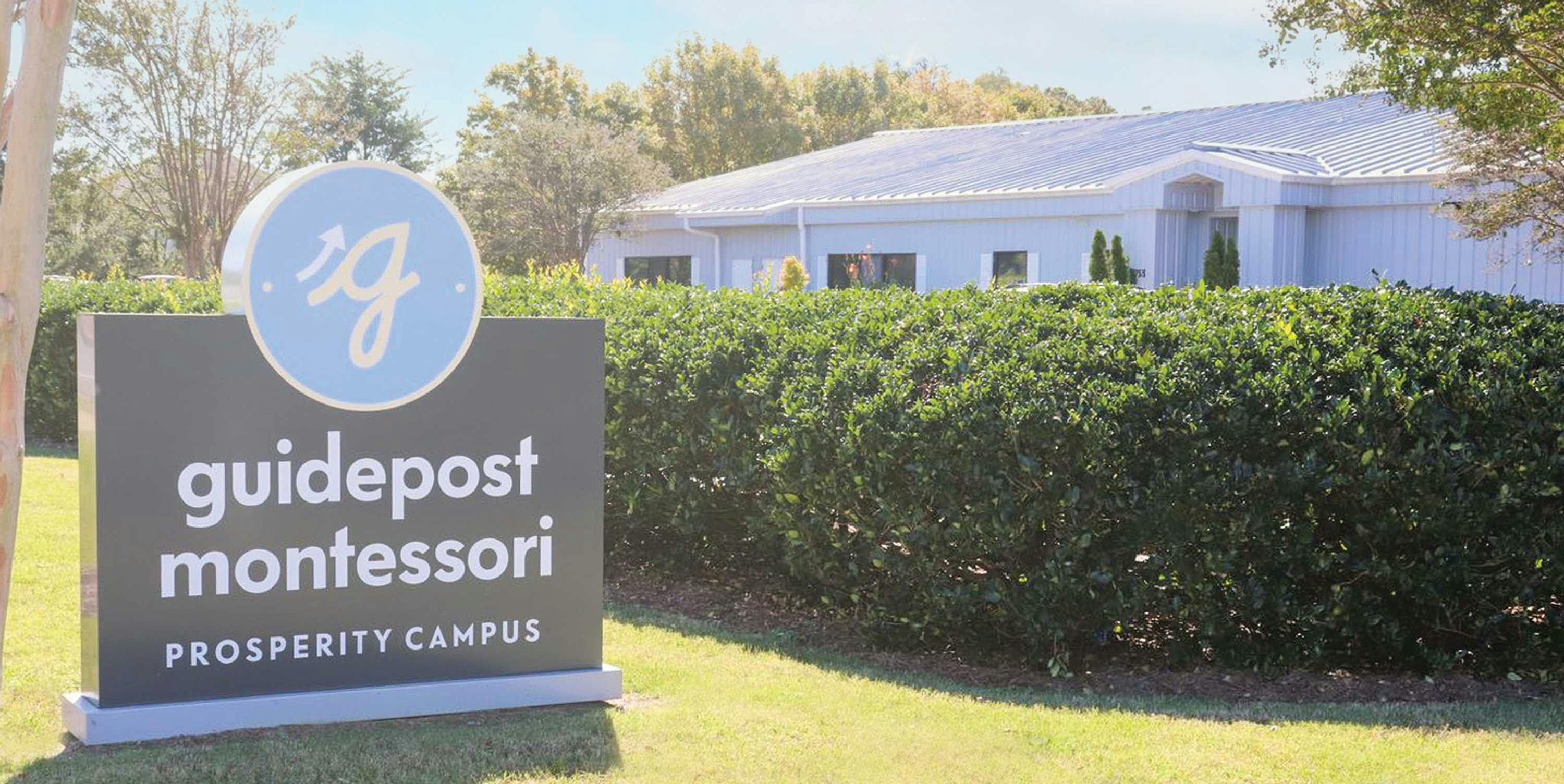 View of Guidepost Montessori at Prosperity from the lawn that shows the top of the campus building over some hedges next to a branded Guidepost Montessori sign that reads "Prosperity Campus"
