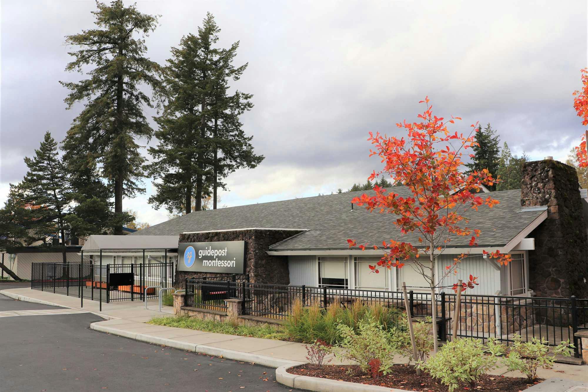 View of Guidepost Montessori at Beaverton from the parking lot showing the fenced in campus with its towering trees in the background, a branded Guidepost Montessori sign, and a tree with red and orange leaves in fall time.