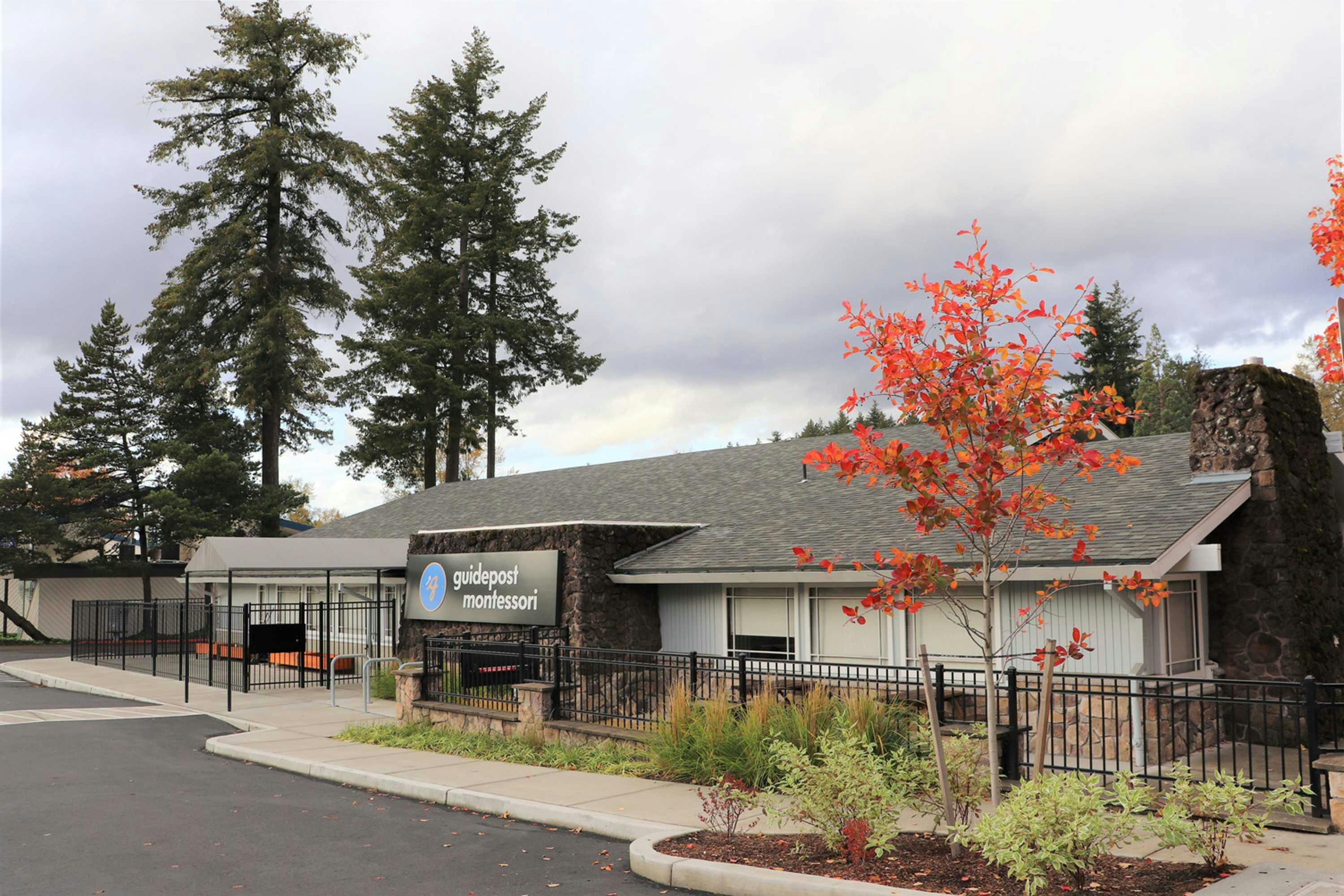 View of Guidepost Montessori at Beaverton from the parking lot showing the fenced in campus with its towering trees in the background, a branded Guidepost Montessori sign, and a tree with red and orange leaves in fall time.