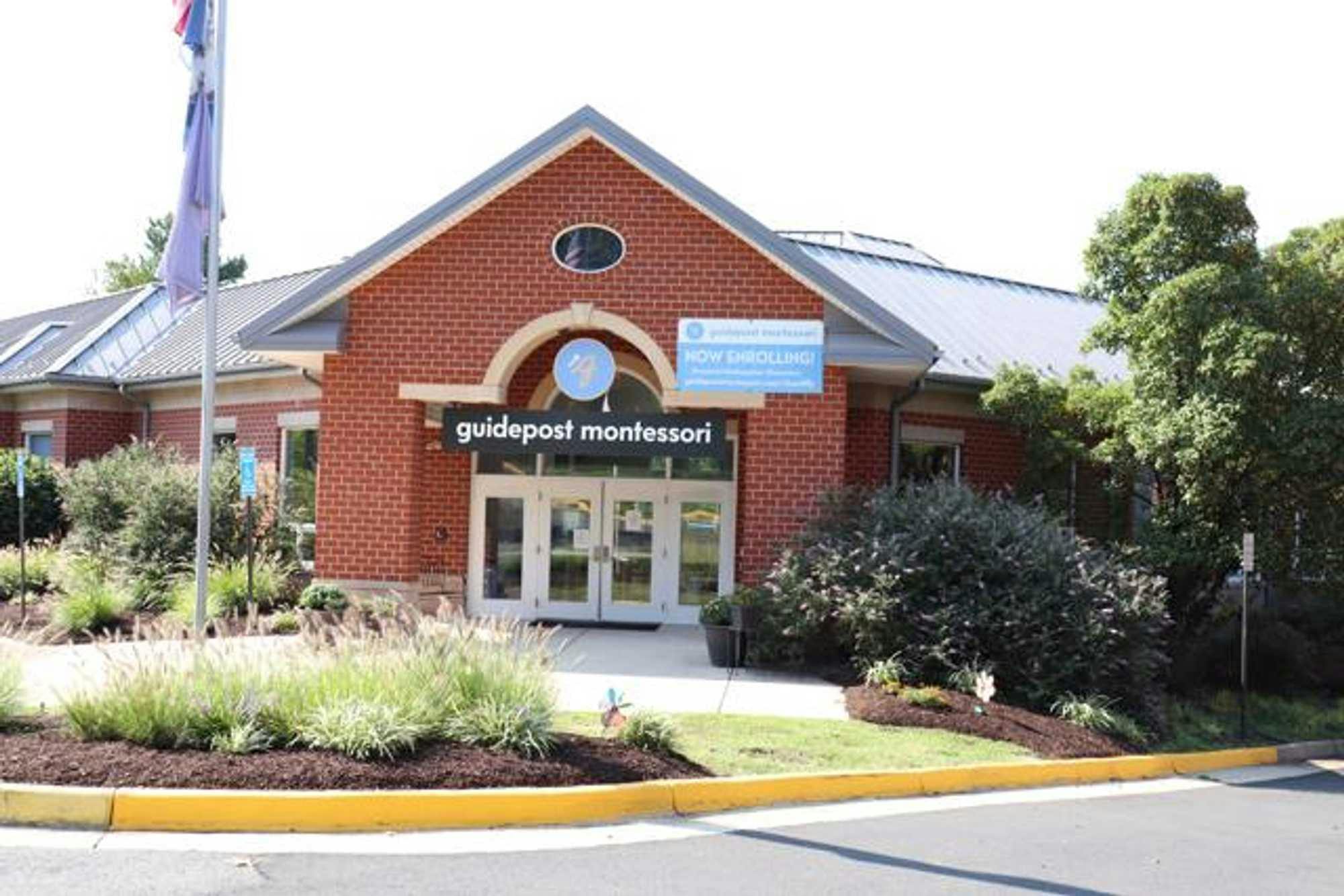 View of Guidepost Montessori at Chantilly from the parking lot that shows the brick exterior and entrance with four glass doors. A branded Guidepost montessori sign hangs above the entrance. There is a flagpole out front as well as plants and shrubbery, Trees with dark green leaves loom off screen on the right hand side. 