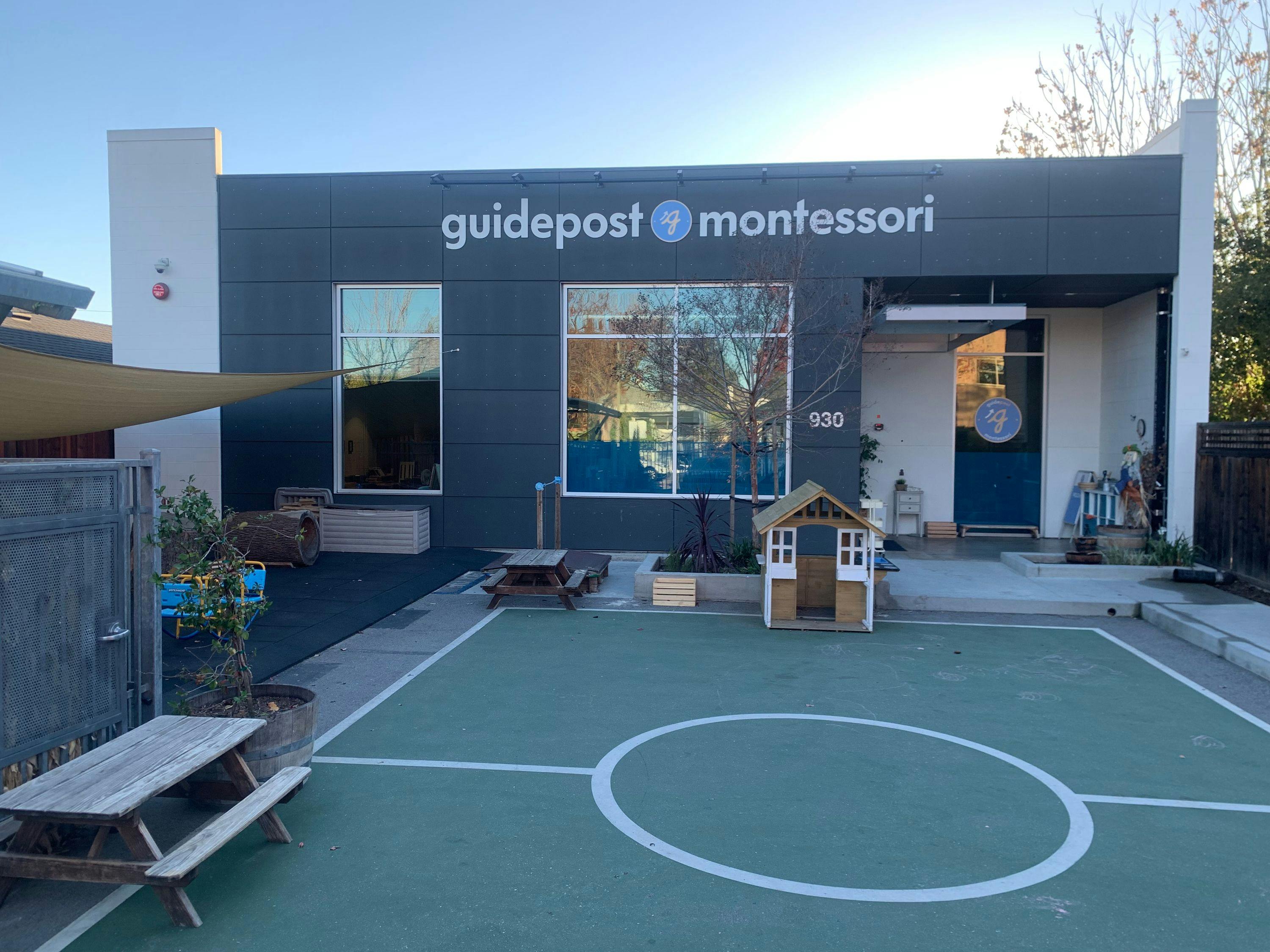 View into the backyard playground of Guidepost Montessori at Palo Alto that shows the gray exterior with a branded Guidepost Montessori sign over outward facing windows.