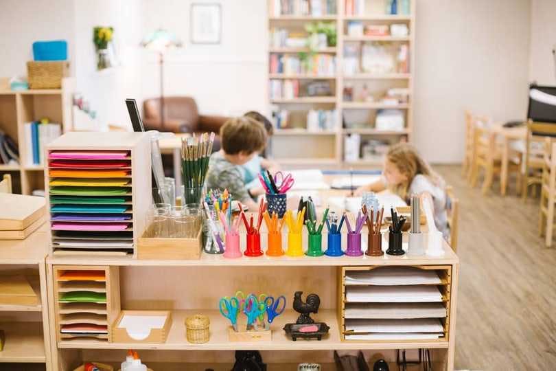 helves are stacked with colorful paper and organized with classroom tools like scissors, pens, paintbrushes and glue. Students complete work in the background. 