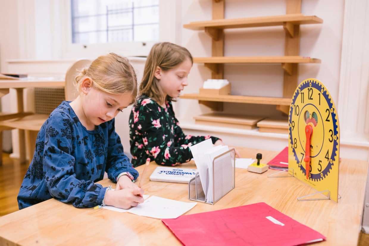two little girls writing on paper at a table next to a large yellow clock