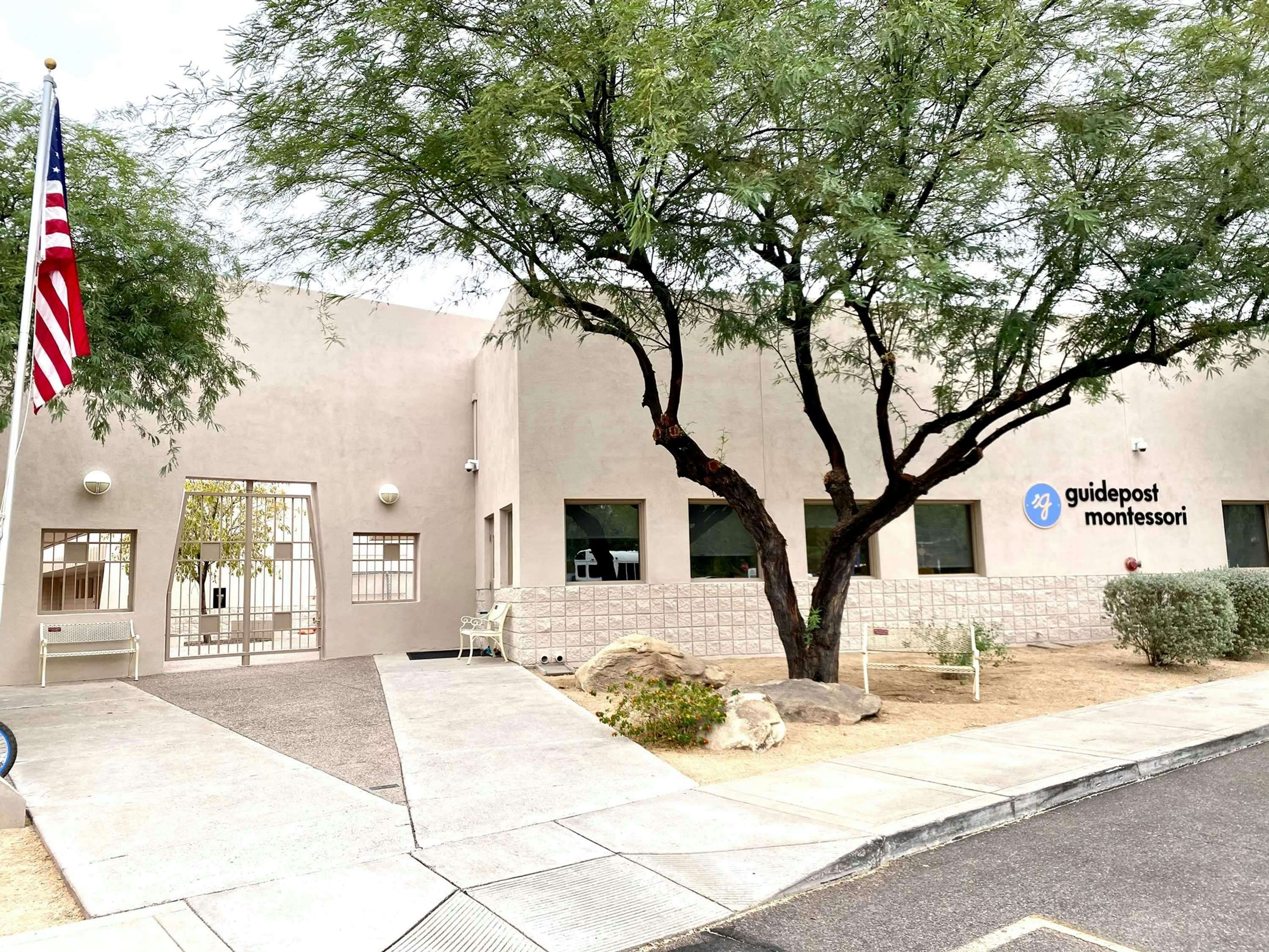 View of Guidepost Montessori at North Scottsdale from the parking lot that shows the entrance gate and a tree in front of four windows with a Guidepost Montessori branded sign on the right facade of the building.