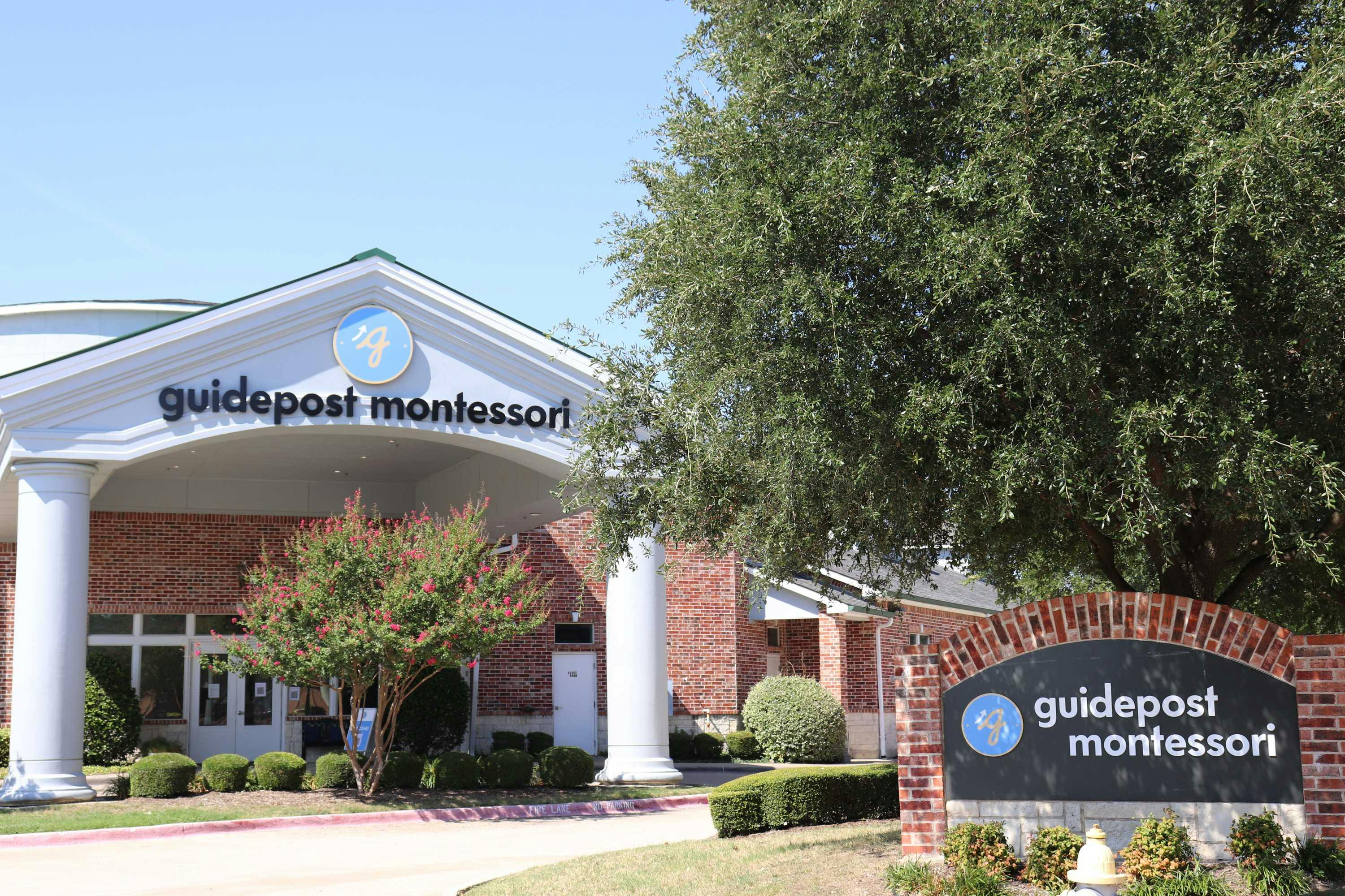 View of Guidepost Montessori at Stonebriar that shows the floral walkway, lined with bushes and blossoming trees, flanked by two large white pillars. Two branded Guidepost Montessori signs can be seen.