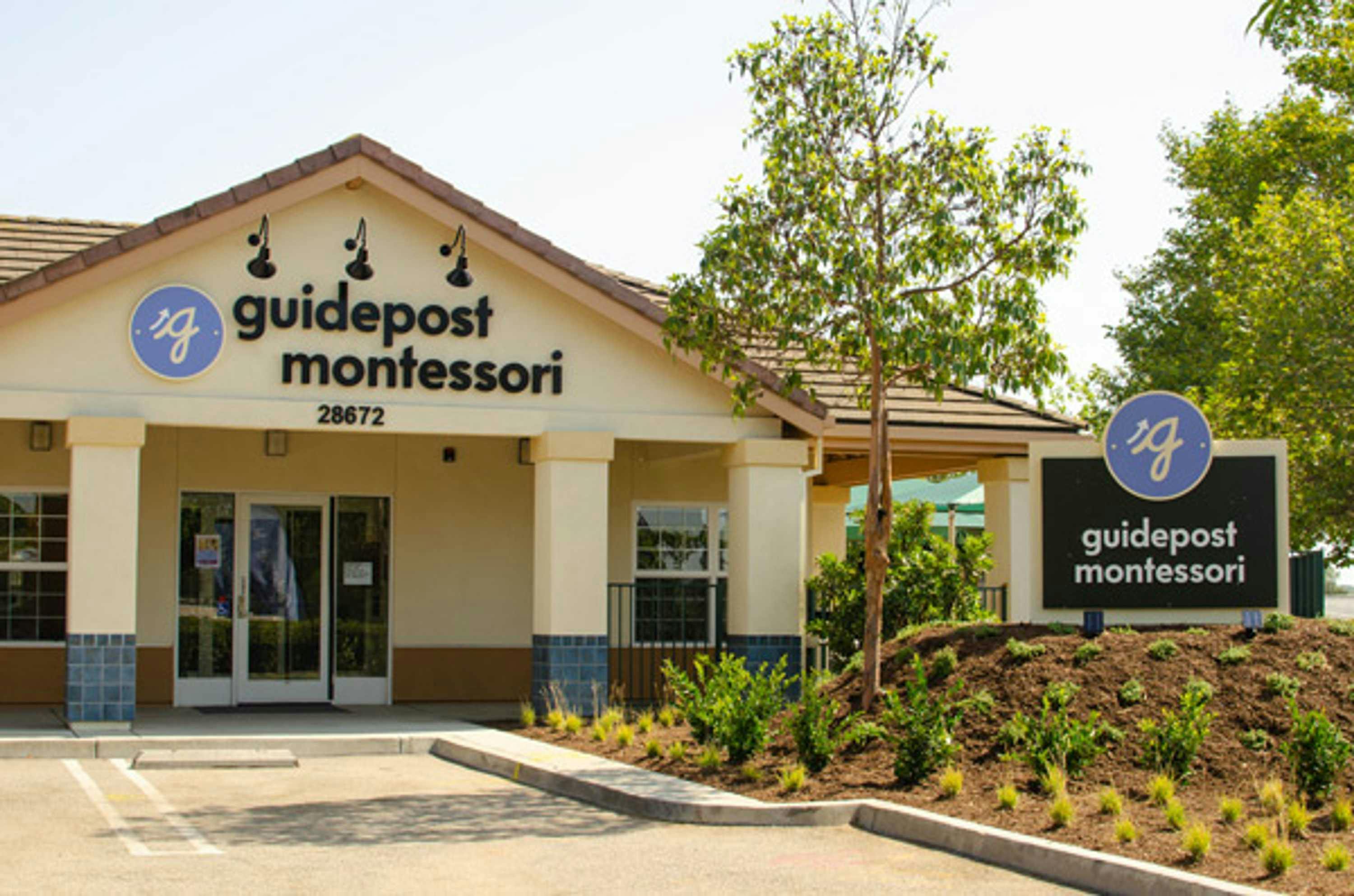 View of Guidepost Montessori at Las Flores entrance from the parking lot that shows the pillared entrance and a branded Guidepost Montessori sign above the door next to a small planter with another branded Guidepost sign.