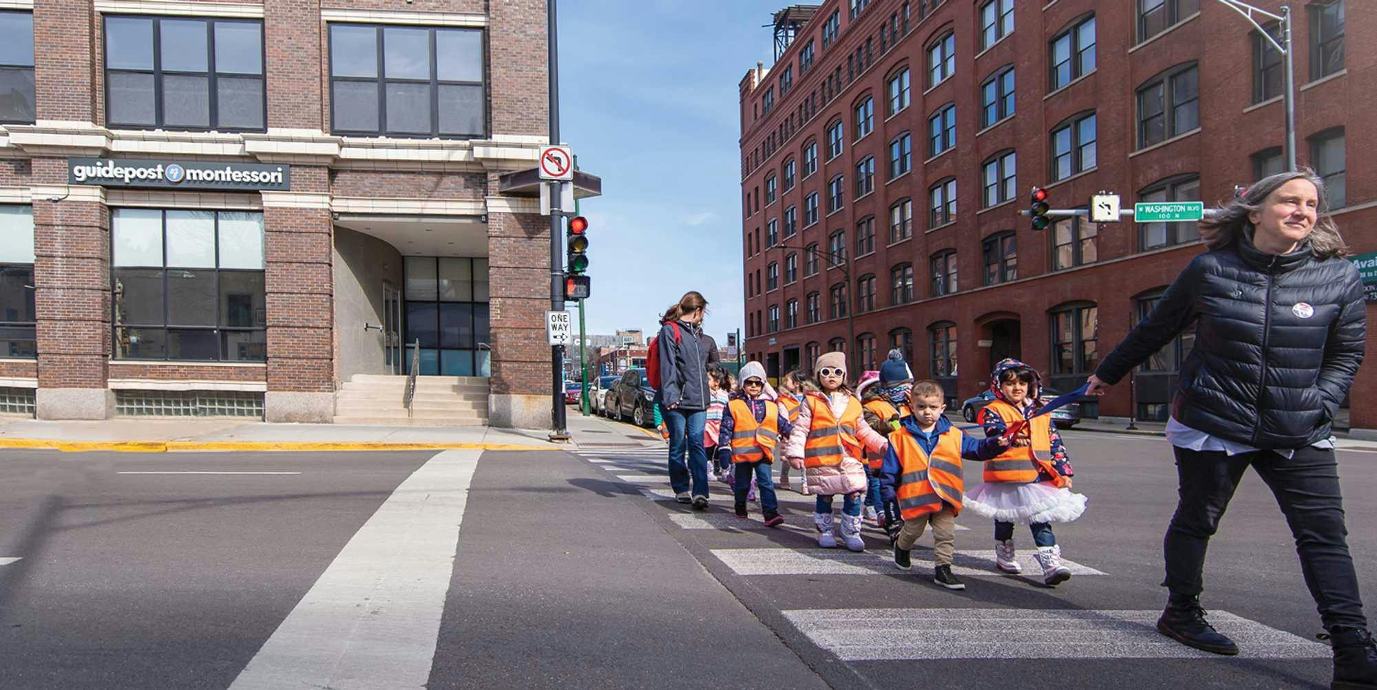 View of Guidepost Montessori at West Loop from the street that shows a class of students in orange safety vests walking in a crosswalk away from the campus. The school is a corner building behind the children and a branded Guidepost Montessori sign can be seen hanging above some windows. Other large buildings and blue skies loom in the background.