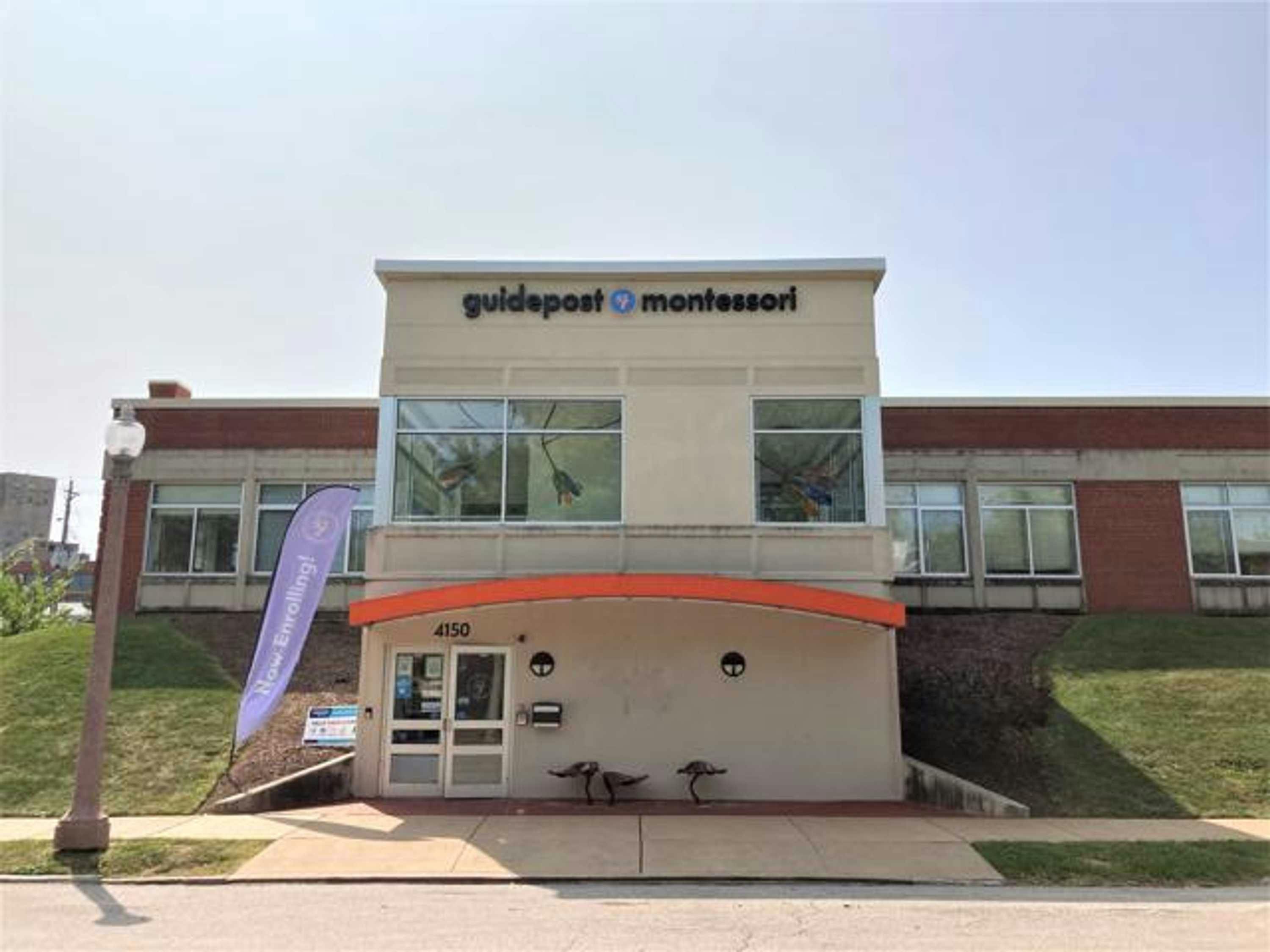 View of Guidepost Montessori at Central West End from the street that shows the exterior of the building with a feather banner in front and an orange awning below a row of windows and a branded Guidepost Montessori sign.