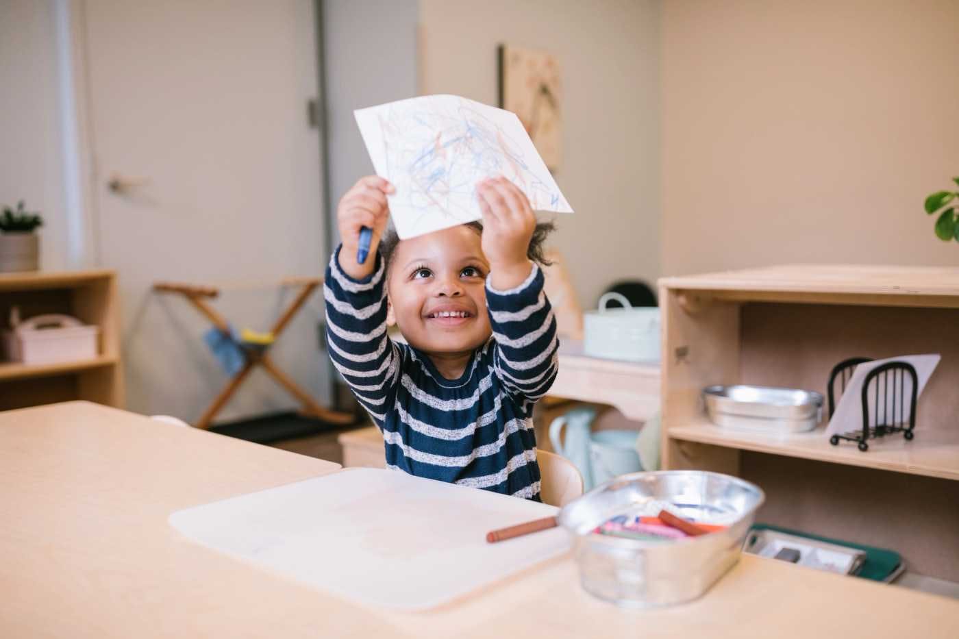 child holding their drawing up over their head while sitting a table in a class room