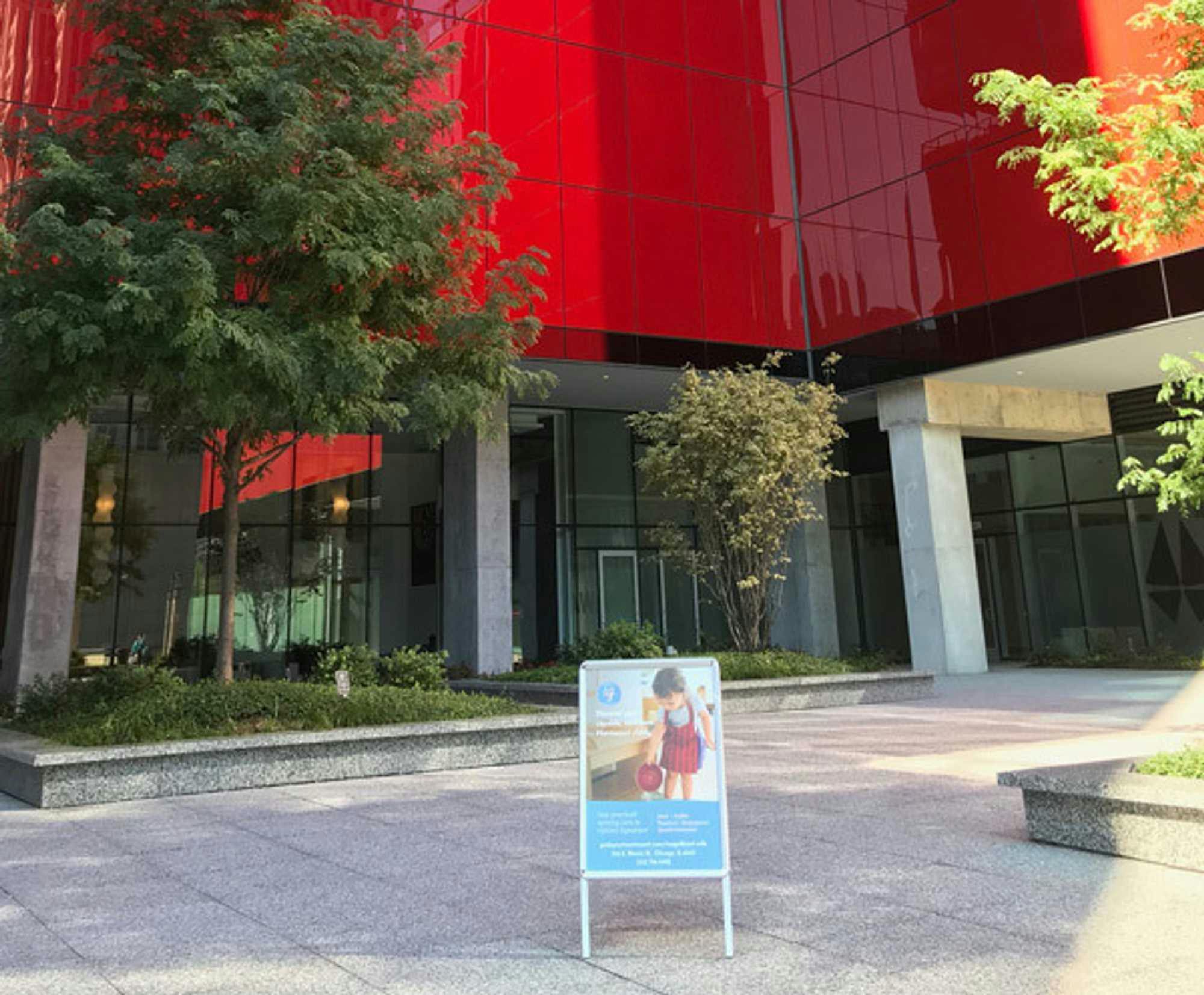 View of Guidepost Montessori at Magnificent Mile showing the towering red skyscraper with trees out front and a branded Guidepost Montessori A-frame sign