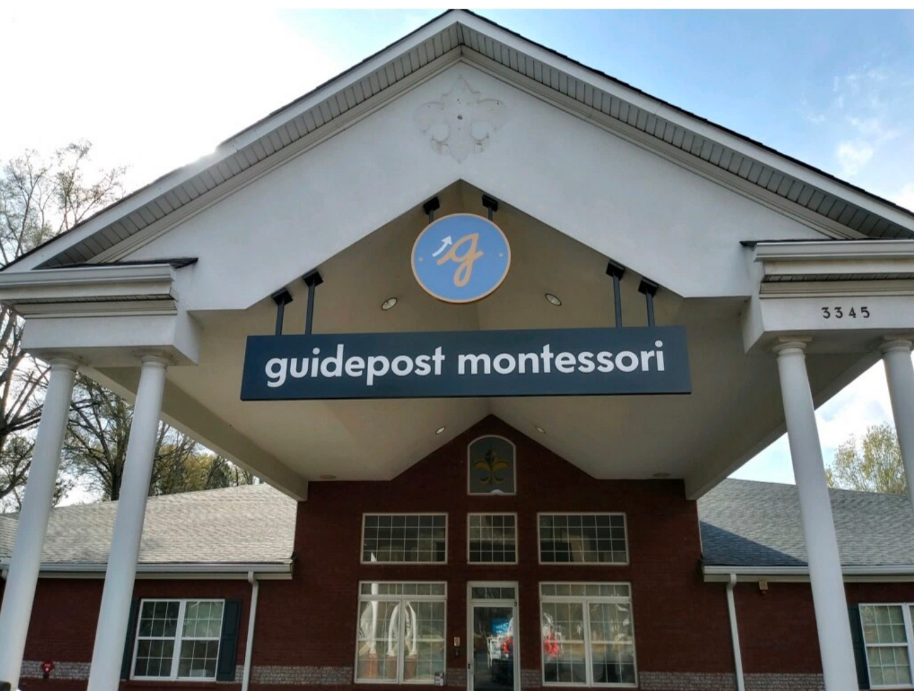 View of the entrance to Guidepost Montessori at Duluth that shows the pillared entryway leading to the glass doors and windows of the entrance. A branded Guidepost Montessori sign hangs from under the overhang.