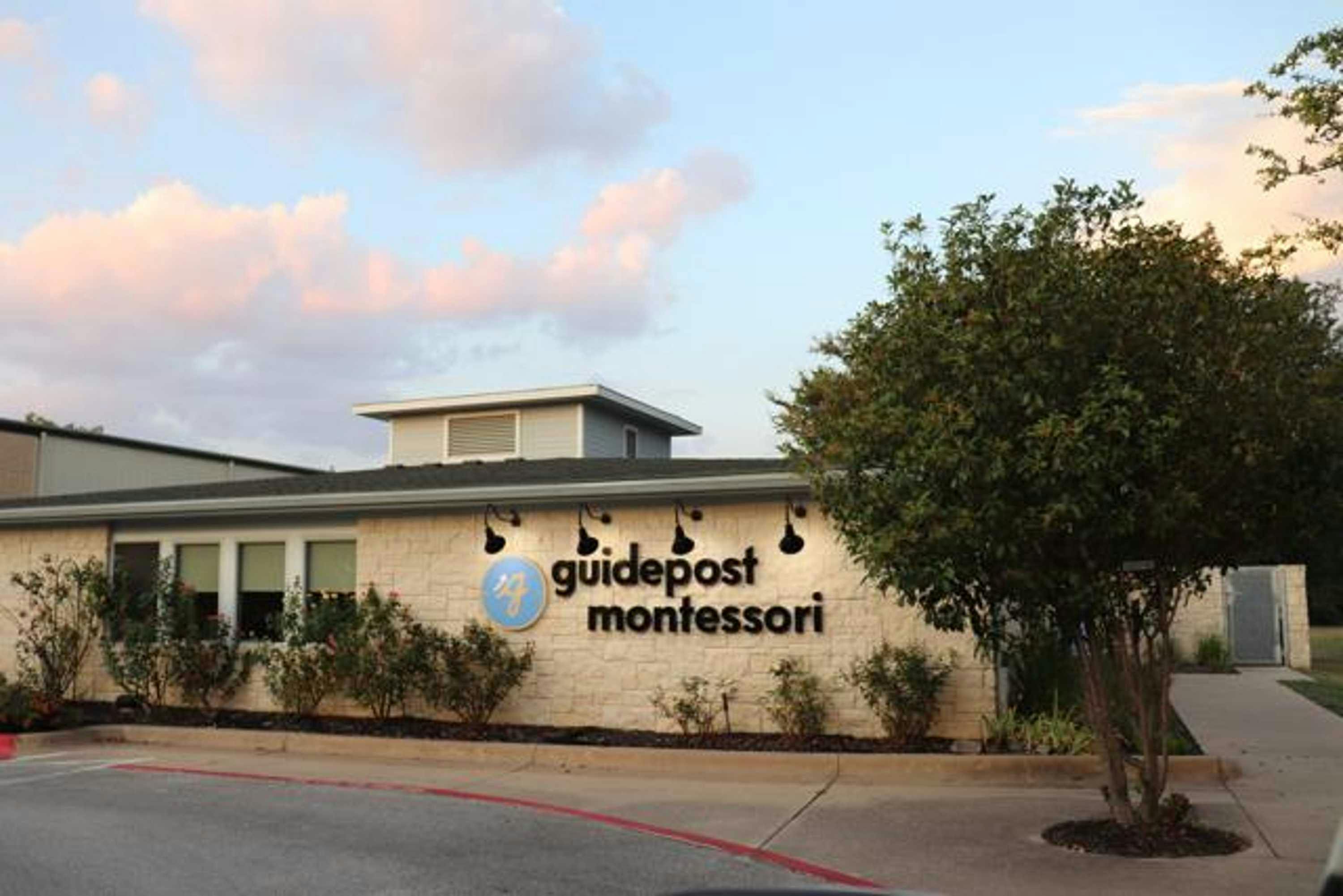 View of Guidepost Montessori at Cypress Creek from the parking lot that shows the block facade of the one-story building with a branded Guidepost Montessori sign. Some bushes line the building and a lone tree stands to the right. A sunset looms in the background.