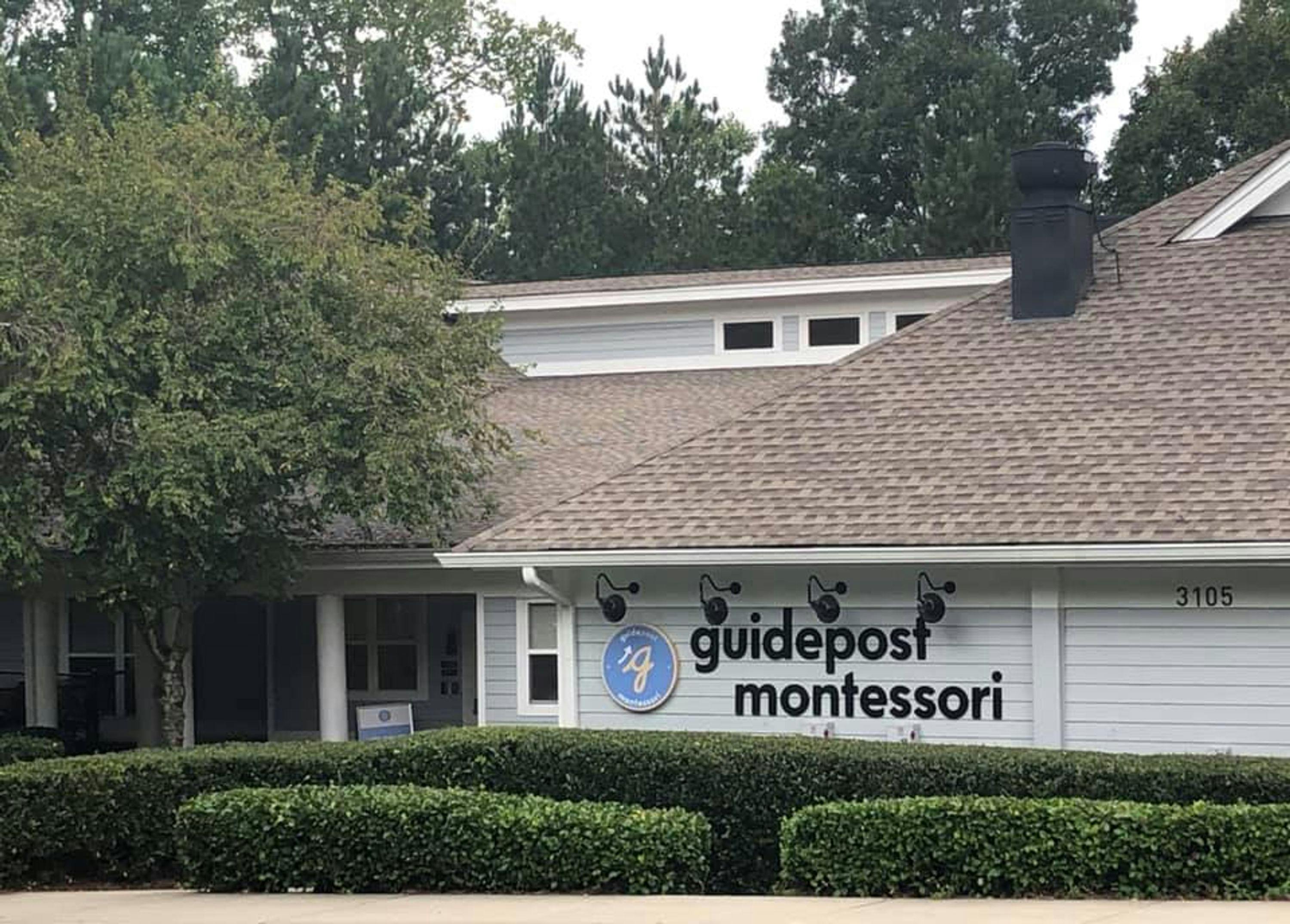 View of Guidepost Montessori at Deerfield through the hedges, showing the closed in walkway shaded by a tree and a branded Guidepost Montessori sign hanging on the right side.