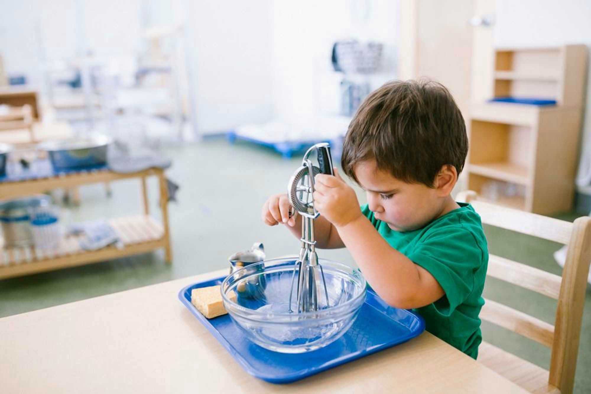 Young boy learns to use a whisk in a clear bowl on top of a blue food tray. He is sitting in a traditional Montessori environment.