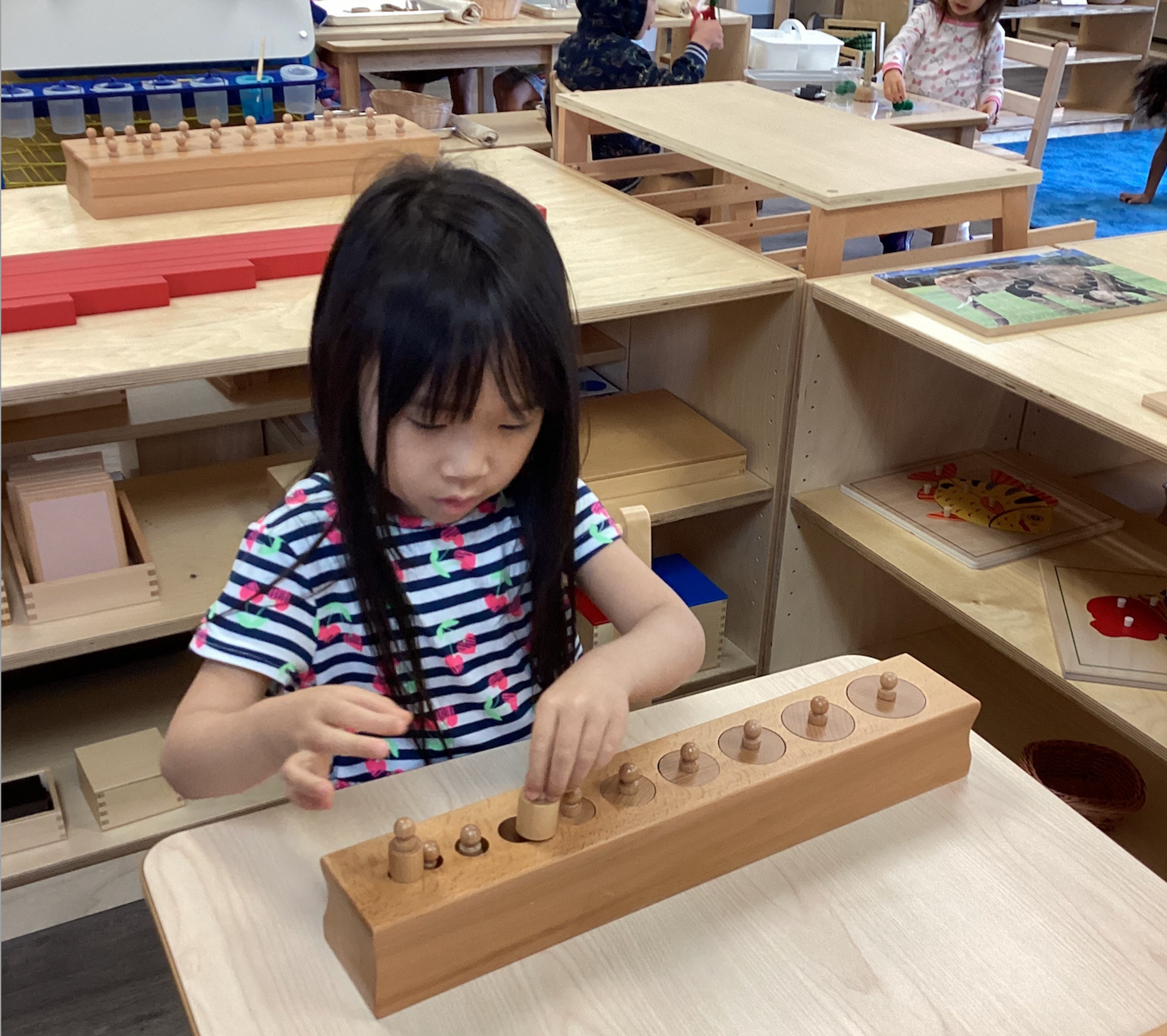 Children in a well-organized mixed-age Montessori classroom ponder over an activity using blocks of different sizes, classic Montessori classroom materials 