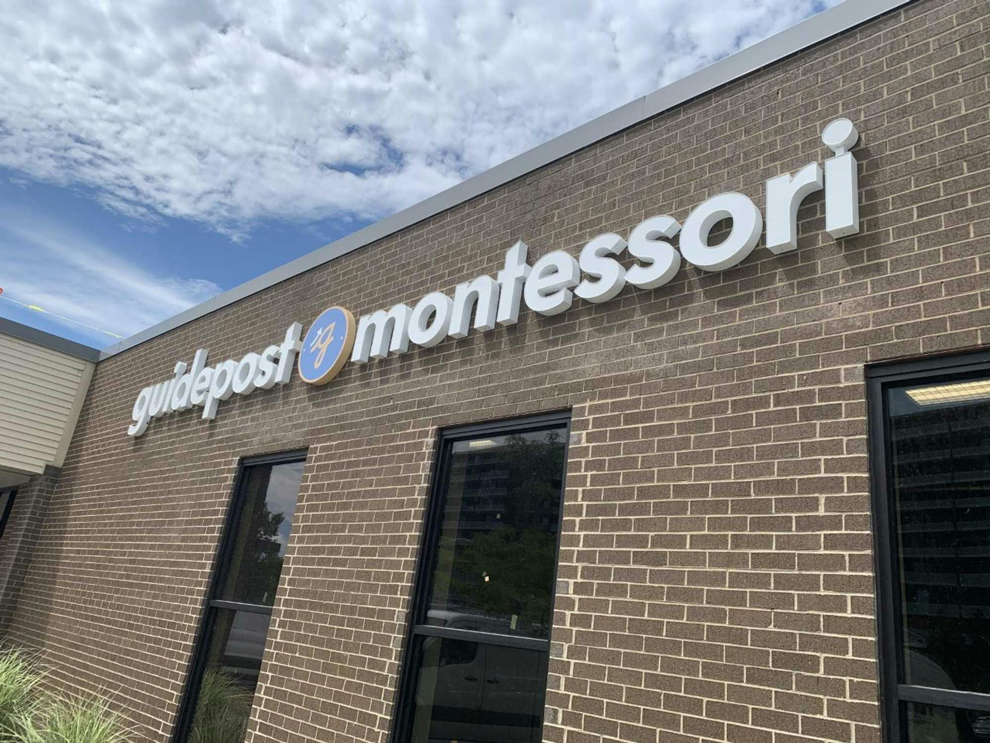Close-Up view of Guidepost Montessori at Schaumburg that shows the brick exterior and 3 exterior windows as well as a branded Guidepost Montessori sign. Blue skies loom in the background.