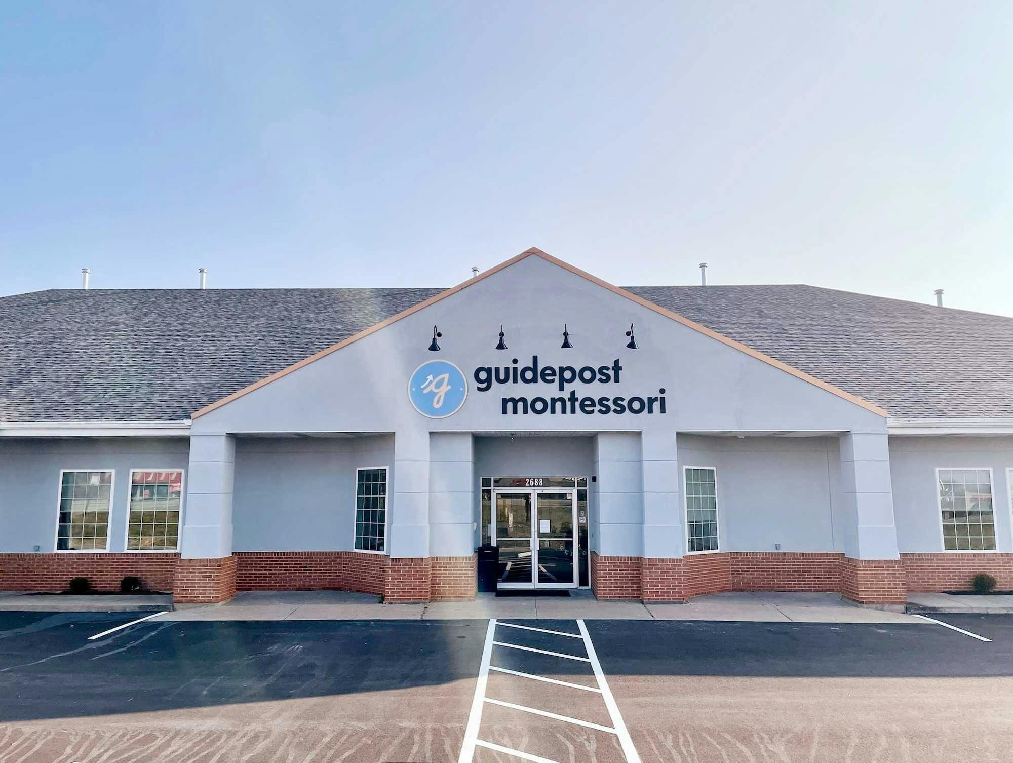 View of Guidepost Montessori at O'Fallon from the parking lot that shows the front exterior of the building with a 4 pillared entrance under a branded Guidepost Montessori Sign