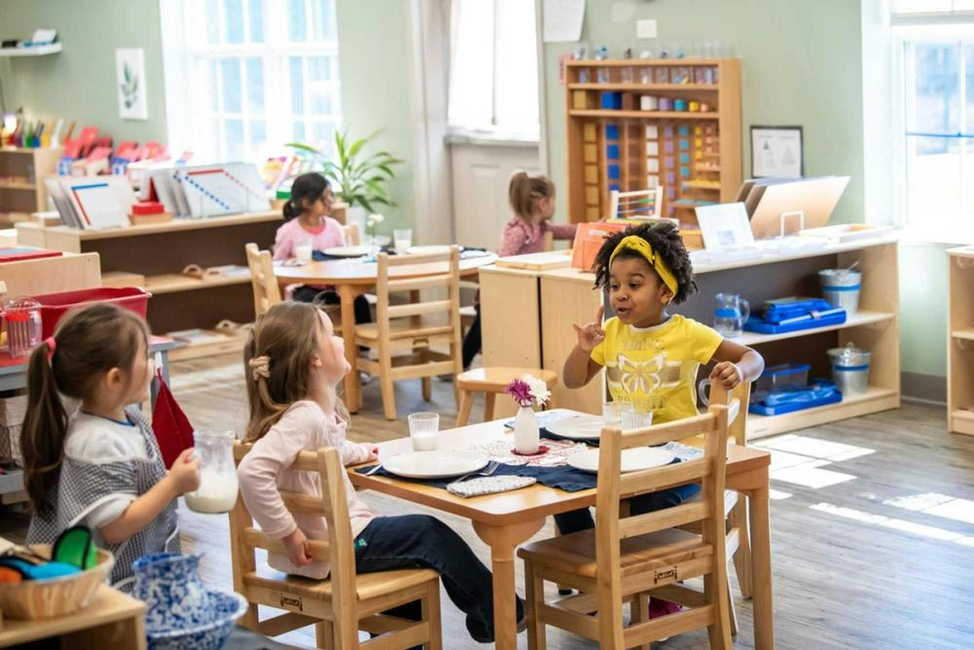 Three kindergarten-aged school girls enjoy a conversation over glasses of milk, while two other students complete activities behind them in the well-organized Montessori classroom