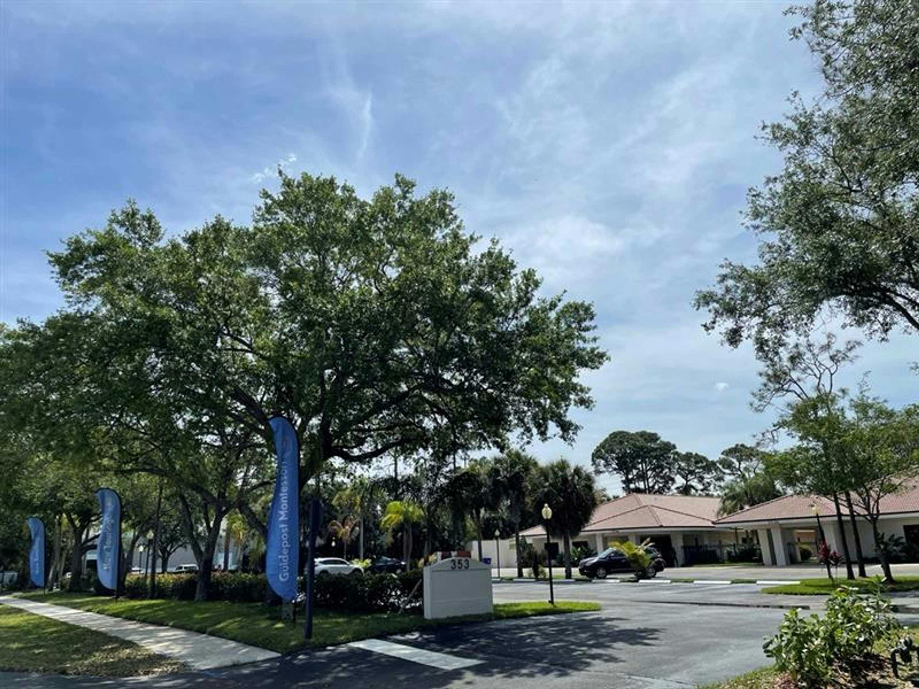 View of Guidepost Montessori at Palm Beach Gardens from the street that shows the shaded parking lot with the campus looming in the background. Branded Guidepost feather banners line the sidewalk to the parking lot entrance.