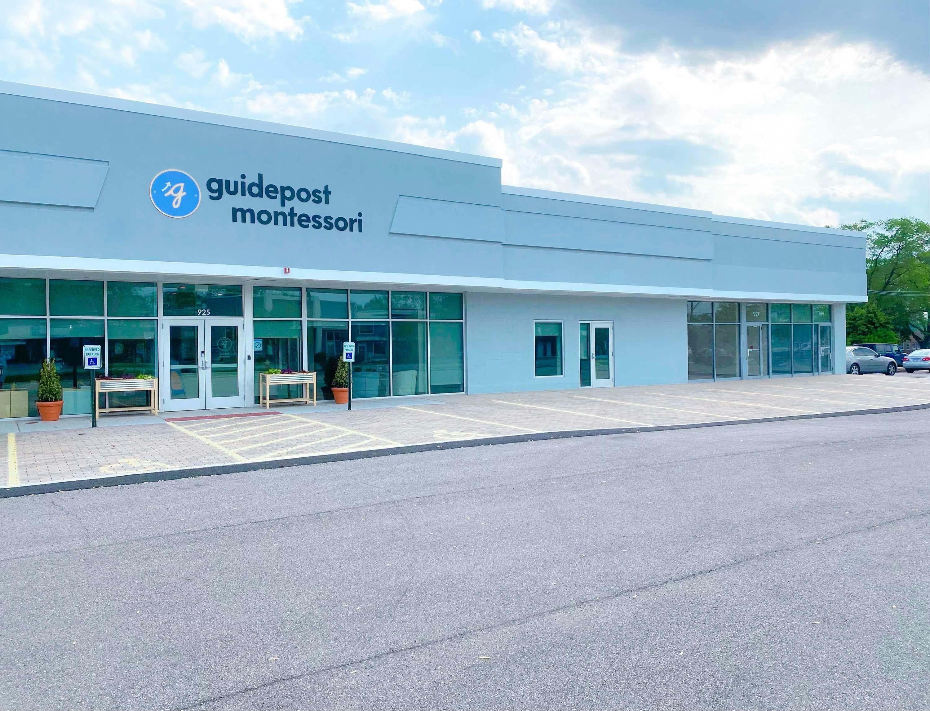View of Guidepost Montessori at Downers Grove from the parking lot that shows the expansive facade of the gray building with a windowed entrance. Some handicap parking spots reside out front and the corner of the building is all windowed. A branded Guidepost Montessori sign sits on the building above the front entrance.