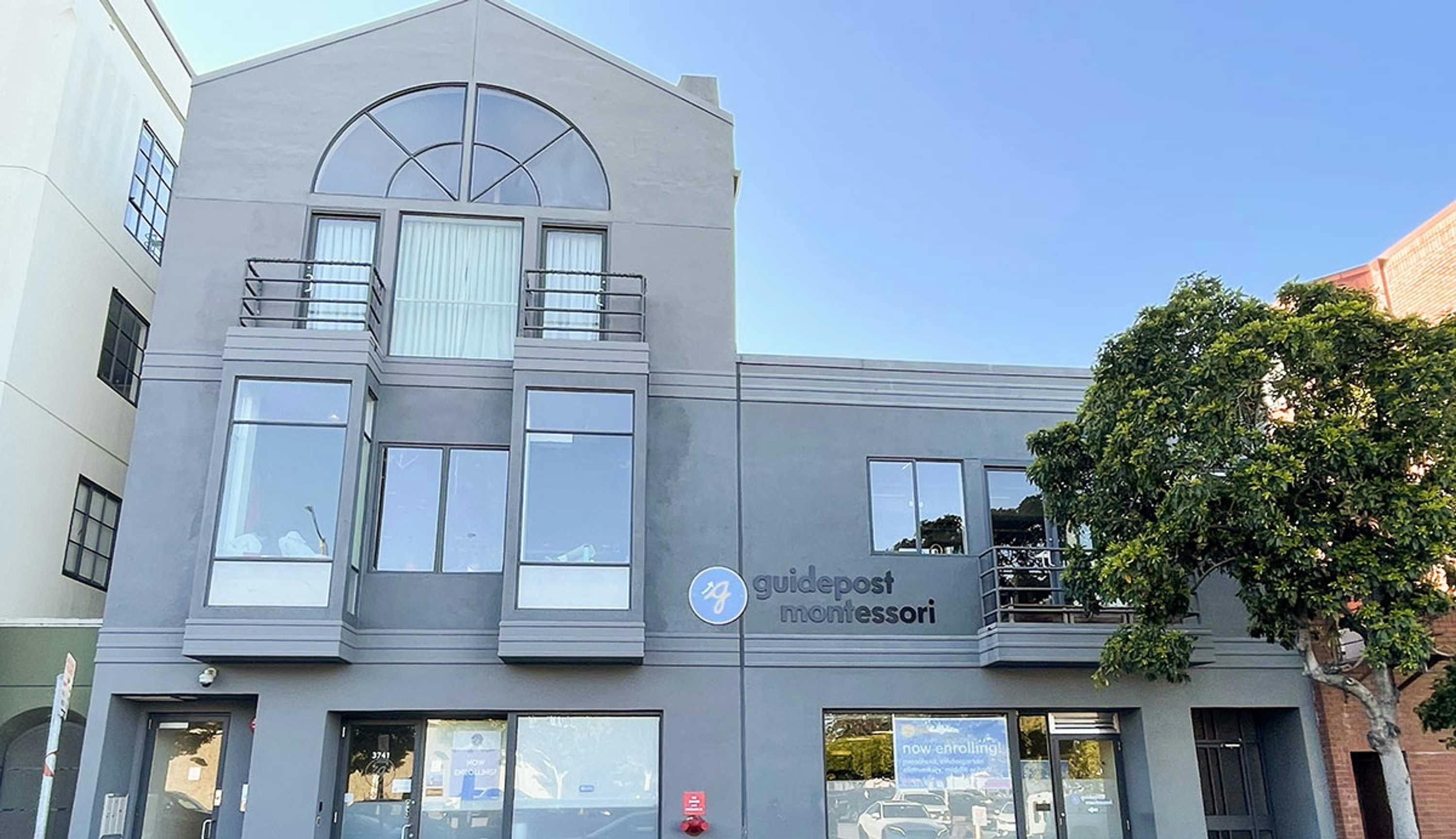 View of Guidepost Montessori at Fort Mason from the street that shows the tall gray, windowed exterior entrance with a large circular Guidepost Montessori branded sign